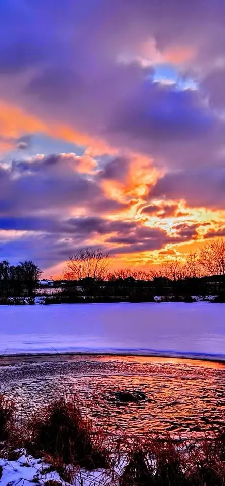 A vibrant, vertically oriented photograph captures a dramatic winter sunset over a frozen body of water. The sky is ablaze…