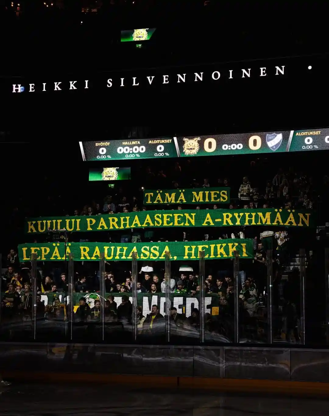 A hockey arena scoreboard shows 'HEIKKI SILVENNÖINEN' above a scoreboard with all zeros. Below, fans hold green banners with…