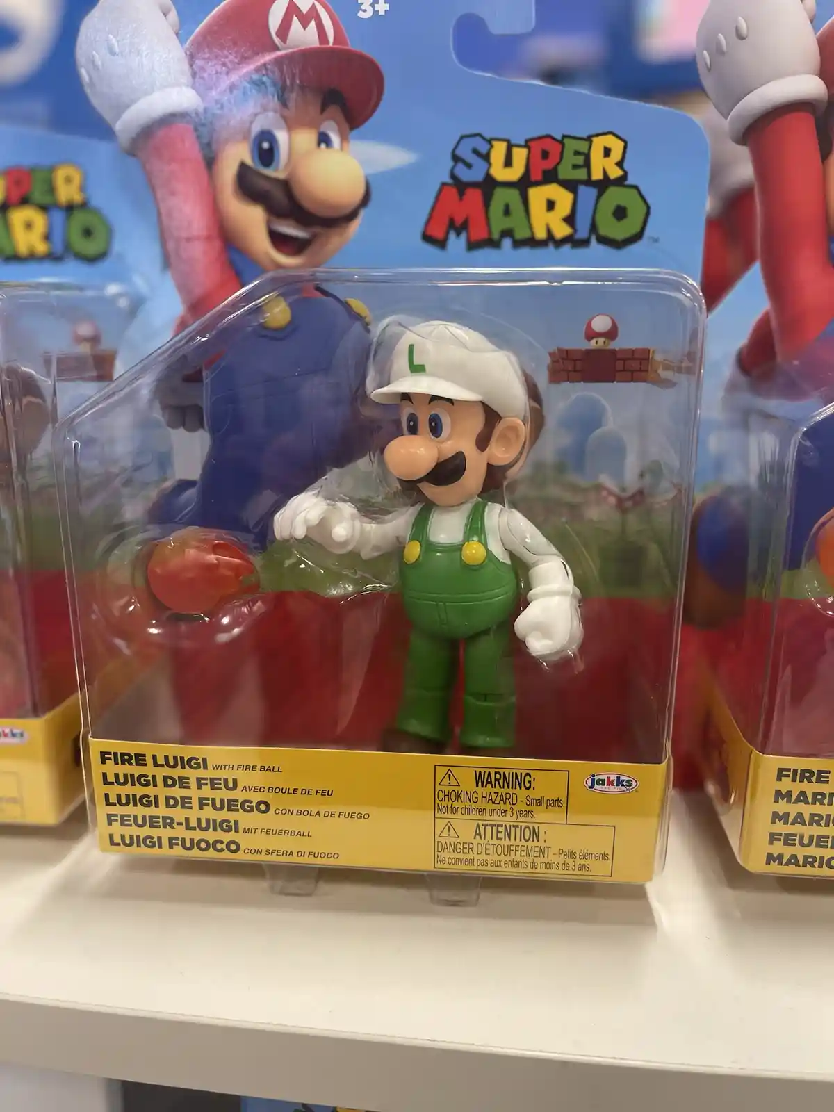 A close-up photo of a Fire Luigi action figure in its retail packaging, displayed on a store shelf. The figure is in his…