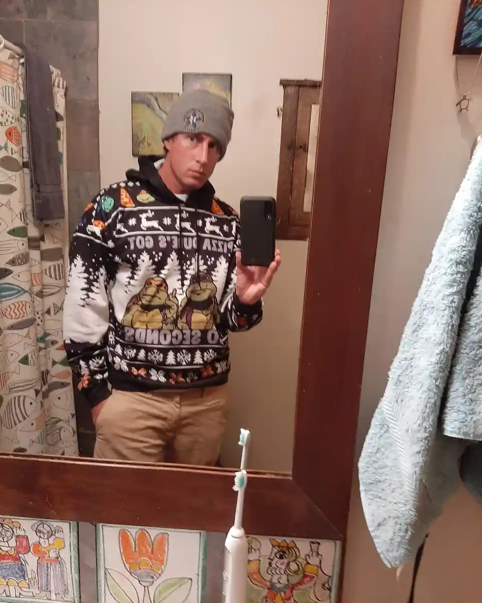 A man in a bathroom mirror selfie wears a black holiday-themed hoodie featuring Teenage Mutant Ninja Turtles and the text…