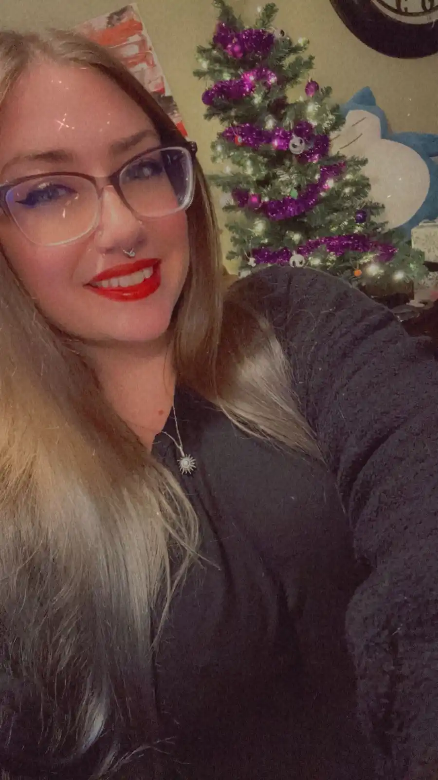 A selfie of a smiling woman with glasses and red lipstick, standing in front of a decorated Christmas tree with purple…