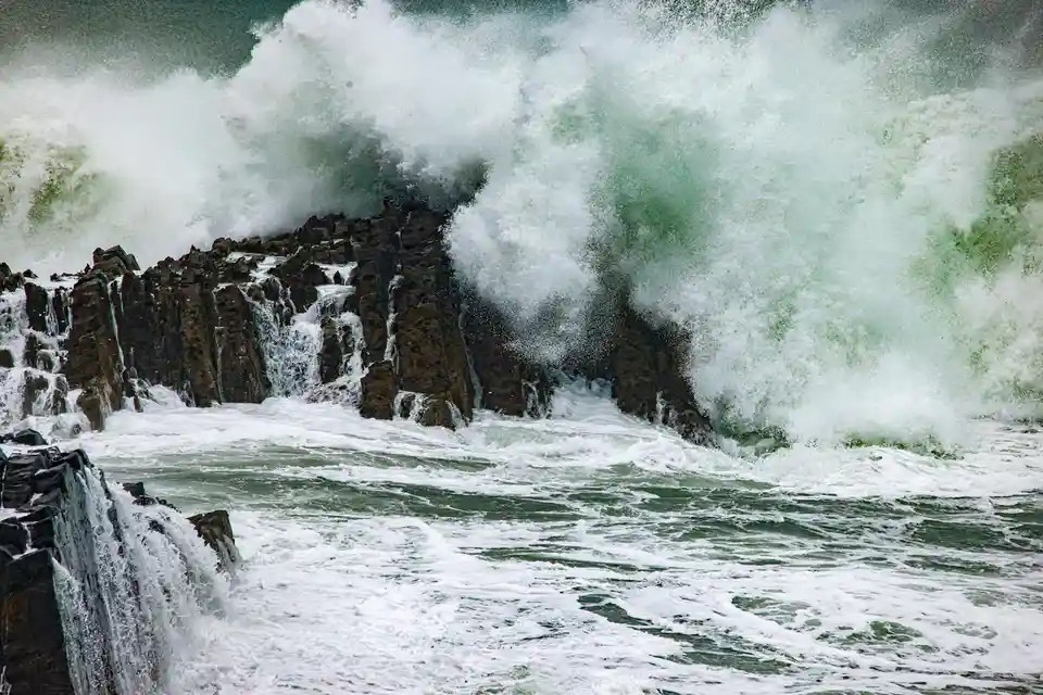 A dramatic photograph shows massive, crashing ocean waves violently splashing against dark, jagged rock formations. The…
