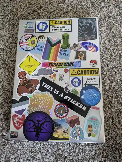 A silver laptop lid is covered in a dense collage of various stickers, including gaming characters like Minecraft's Creeper…
