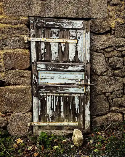 A weathered, peeling blue wooden door set in a rough stone wall, secured with rusty metal bars and a padlock, with grass and…