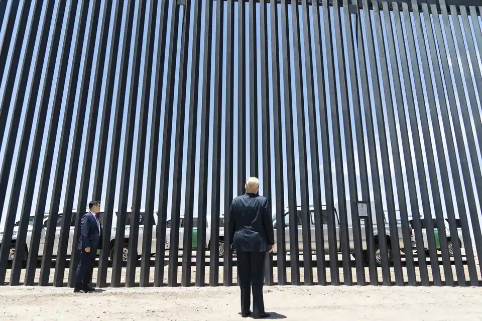A man in a dark suit stands with his back to the camera, facing a tall black metal border fence. Another man in a suit…