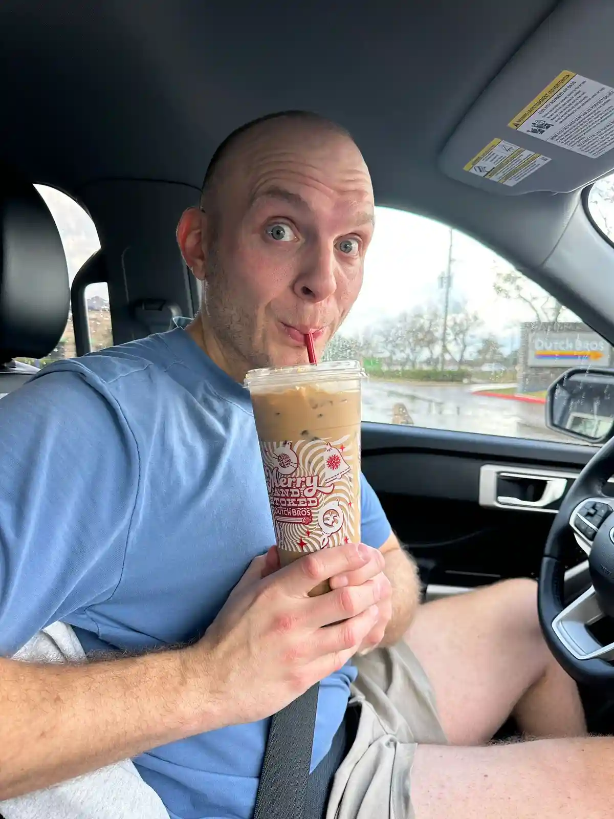 A man with a shaved head and wide-eyed expression sits in the driver's seat of a car, sipping an iced coffee drink from a…