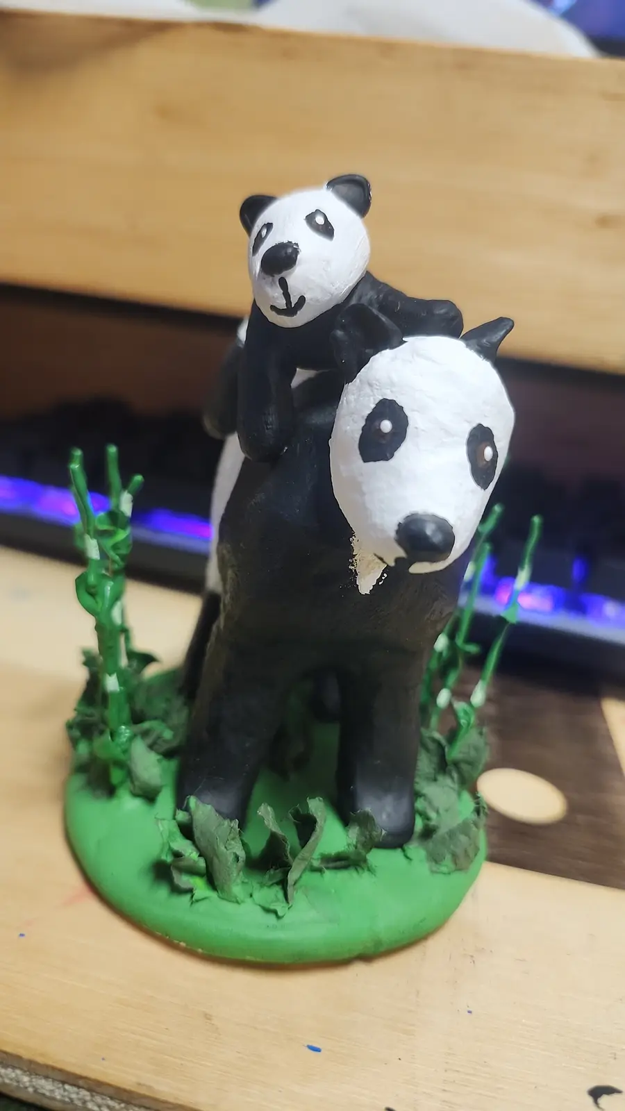 A handmade figurine shows one panda figurine riding on the back of another panda figurine, both standing on a green base…