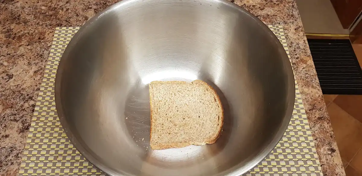 A single slice of whole wheat bread sits alone in the center of a large, empty stainless steel mixing bowl. The bowl rests…