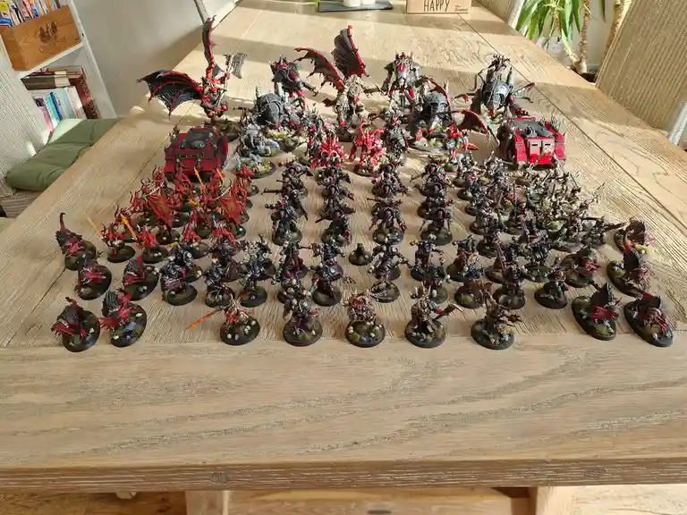 A large wooden table is covered with dozens of intricately painted miniature figurines, mostly in red and black, depicting…