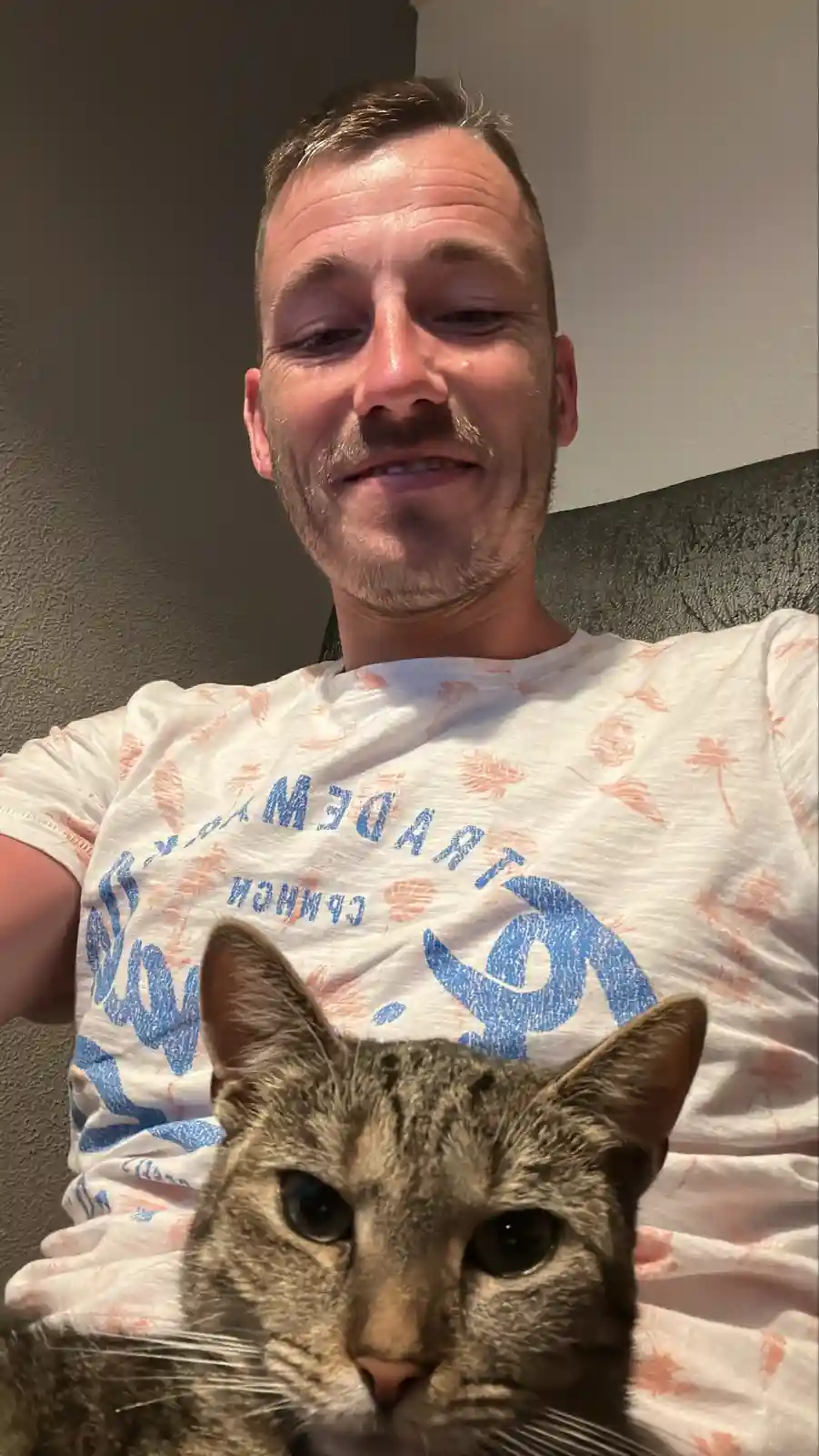 A man with a slightly tearful expression and a mustache is taking a selfie while holding a tabby cat in front of him. The…