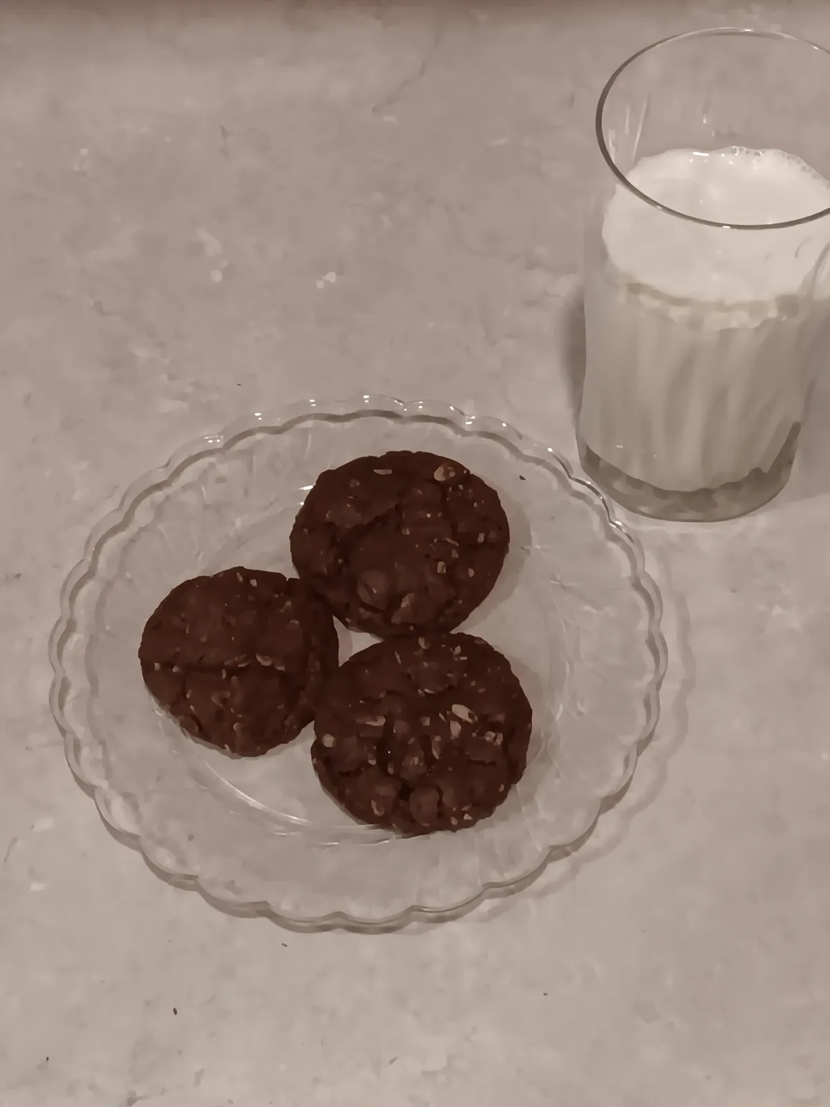 A clear glass plate holds three dark chocolate chip cookies on a light-colored countertop, next to a glass of milk. The…