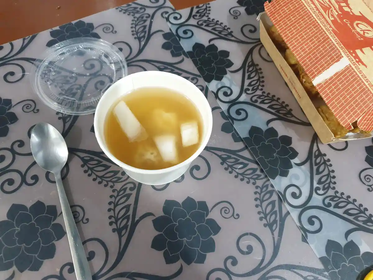 A top-down photo shows a white cup of clear broth with dumplings and a plastic lid beside it, next to a partially open…