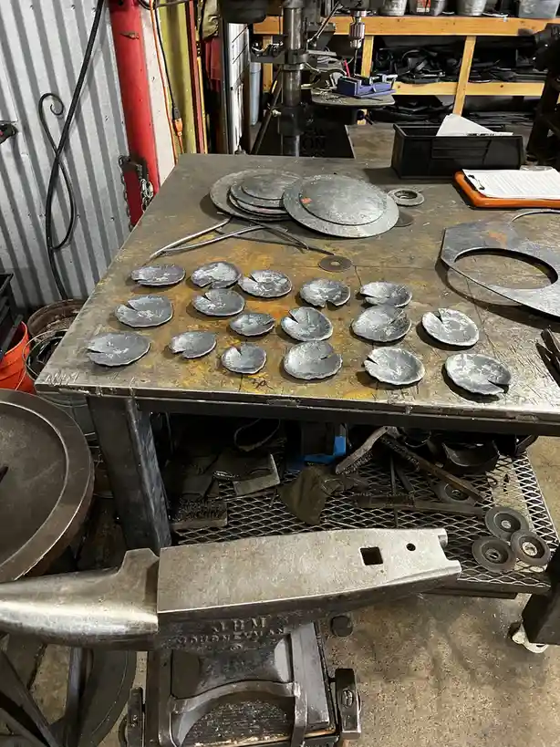 A cluttered metal workshop workbench holds numerous small, circular metal pieces with a single notch, resembling lily pads…