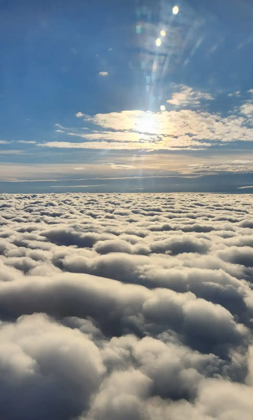 A serene aerial view from above a vast, textured sea of fluffy white clouds under a bright blue sky with the sun shining…