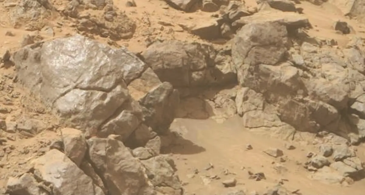 A blurry, reddish-brown rocky landscape, likely from a Mars rover photo, shows large boulders and uneven terrain. Overlaid…