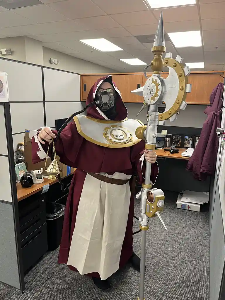 A person in a detailed, full-body costume resembling a grim reaper or inquisitor from the Warhammer 40,000 universe stands…