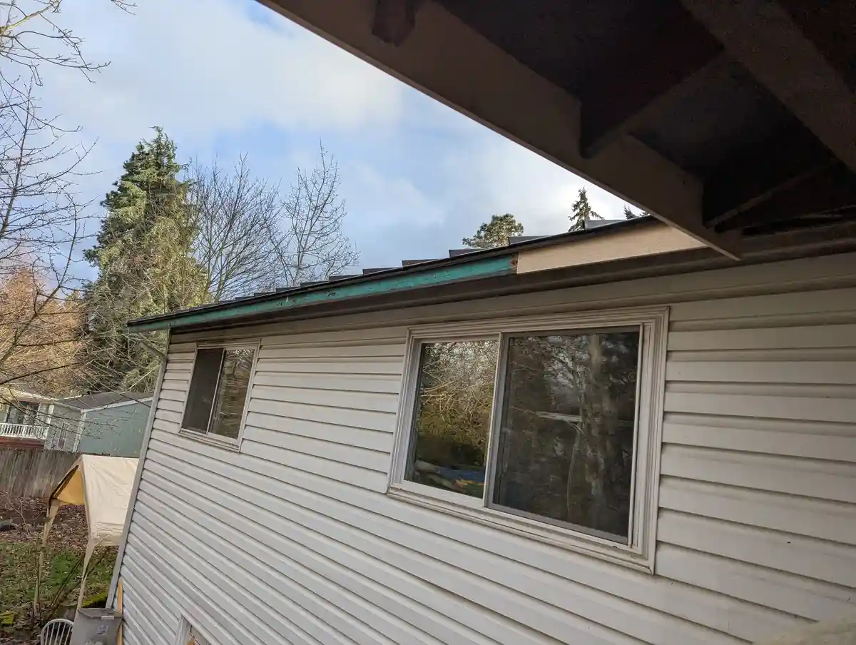 A low-angle photo shows the side of a white-sided house with a porch roof overhead, revealing a slightly damaged or uneven…