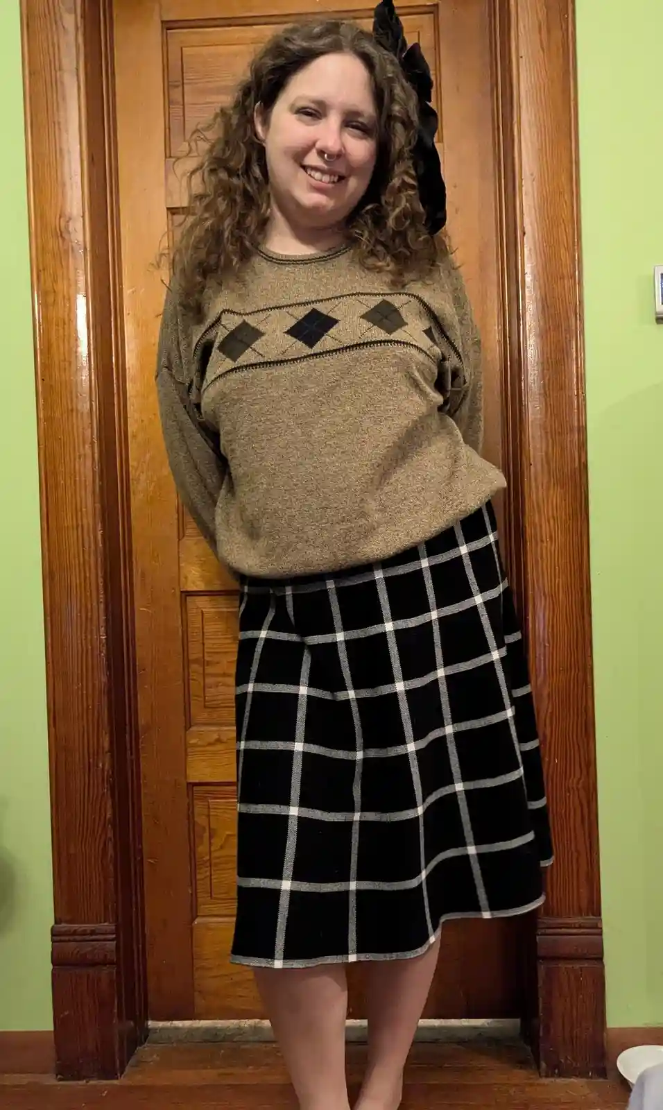 a person with curly hair and a nose ring stands smiling in front of a wooden door, wearing a brown sweater with a black…