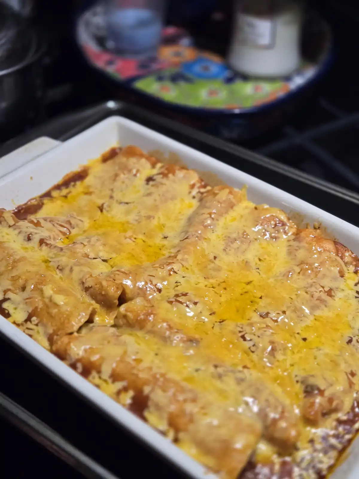 A close-up photo of a white baking dish filled with cheesy enchiladas, with melted, bubbly cheese covering the top. The dish…