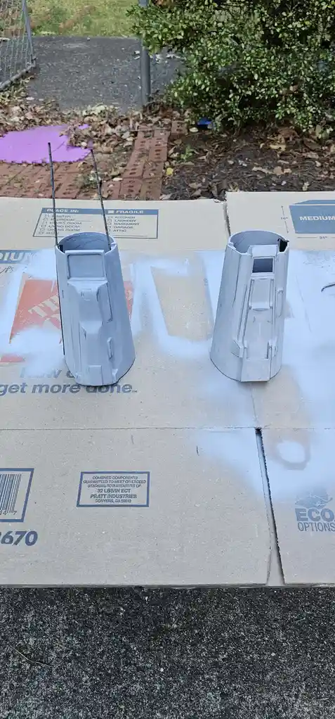 Two gray, angular, sci-fi-style helmet-like objects sit on a cardboard box outdoors, each with thin antennae sticking out…