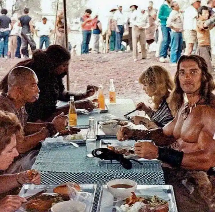 A group of people, including Arnold Schwarzenegger, are seated at a long table eating a meal outdoors. The table is covered…