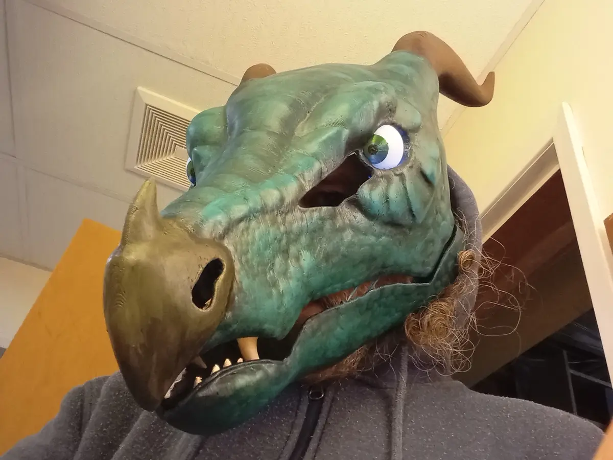 A person wearing a large, green, cartoonish dragon mask with horns, a golden beak-like snout, and big blue eyes, taking a…