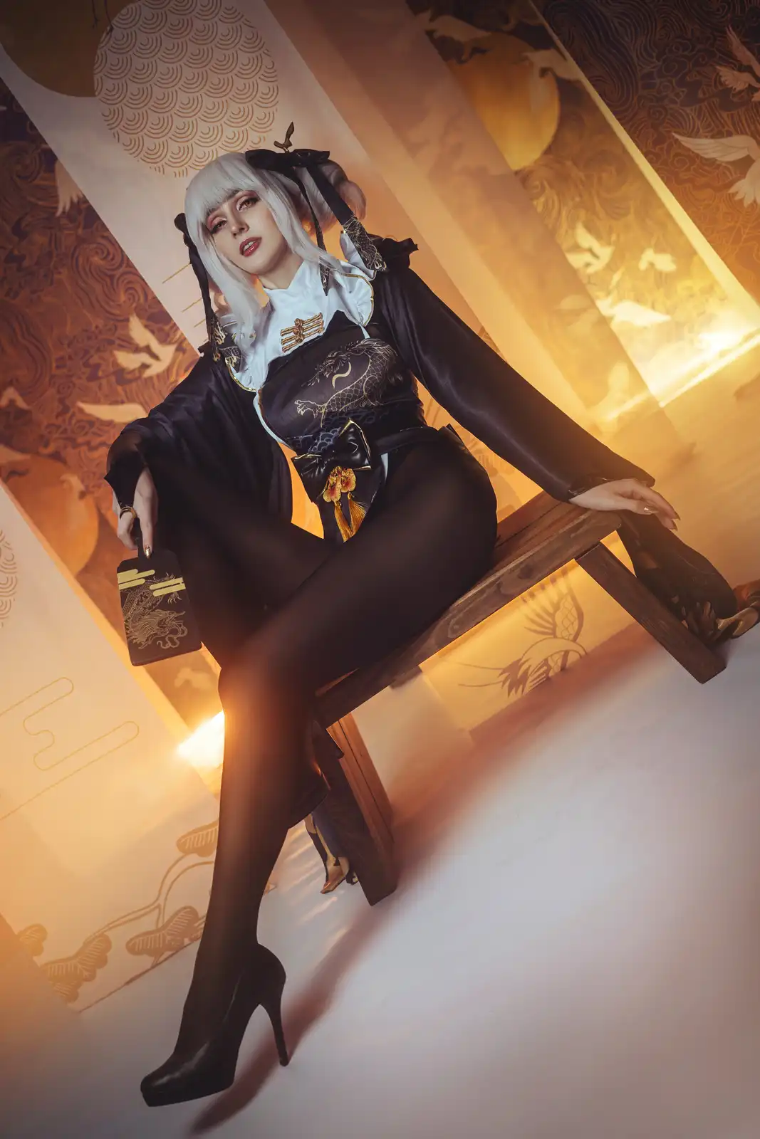 A person with white hair and black bow accessories sits on a wooden bench wearing a black and gold traditional-style outfit…
