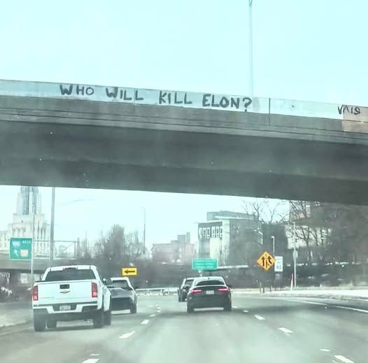 A photograph taken from inside a moving car shows a concrete overpass with the phrase 'WHO WILL KILL ELON?' spray-painted in…