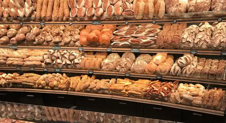 A bakery display case filled with numerous varieties of bread, including baguettes, rolls, sourdough, and pretzels, arranged…