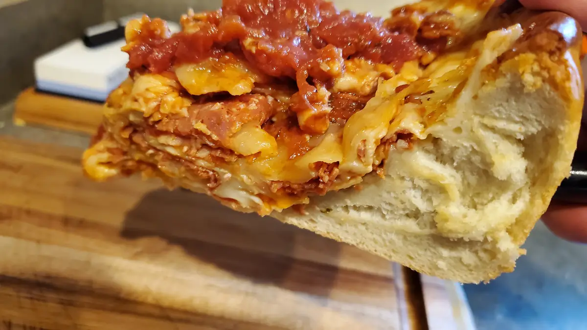 A close-up photo shows a hand holding a thick sandwich with a soft, porous bread roll. Inside, multiple layers of pasta…