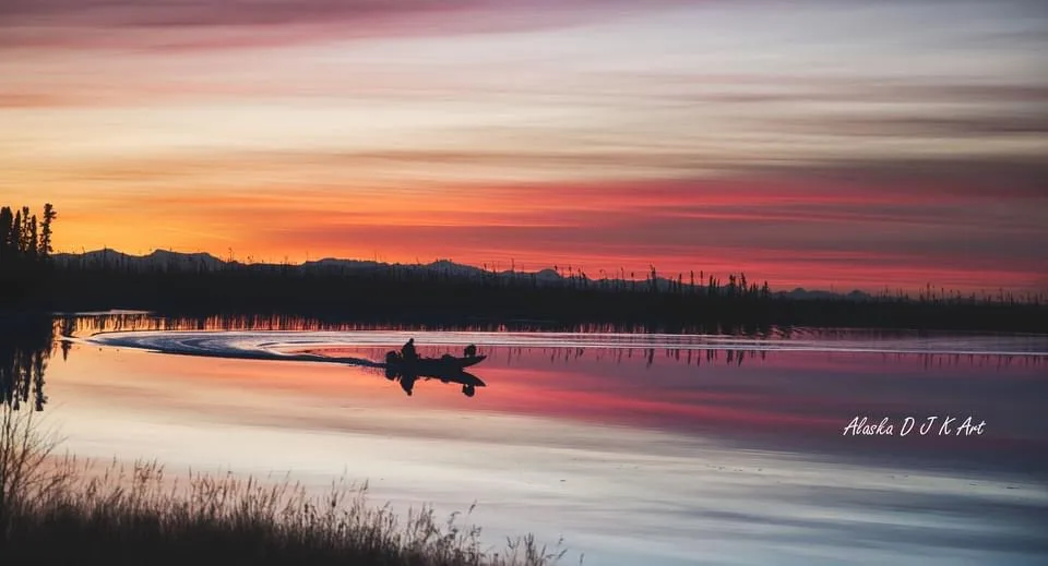 A serene sunset over a calm Alaskan lake, with a person in a small boat leaving a wake. The sky is streaked with vibrant…