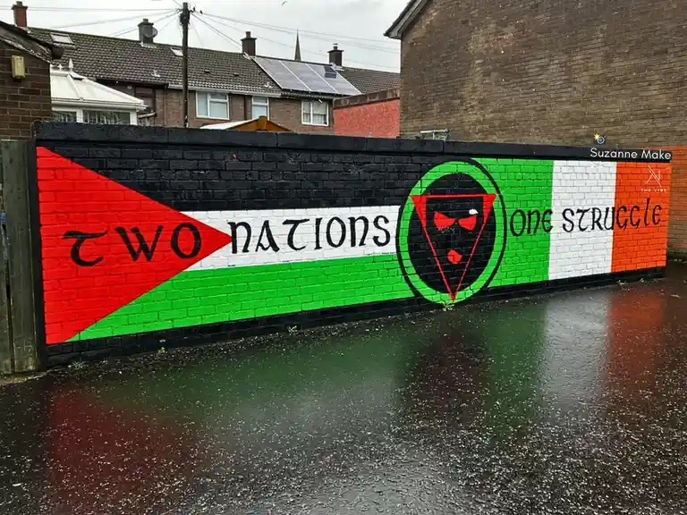 A brick wall mural painted with the Palestinian flag on the left and the Irish tricolor on the right, separated by a central…