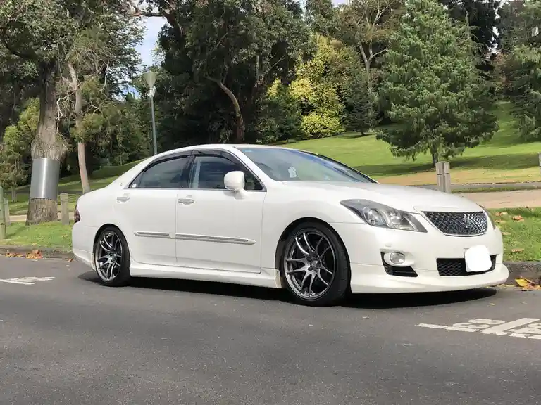 a white toyota mark ii sedan is parked on an asphalt road in a park with green grass and trees in the background. The car…
