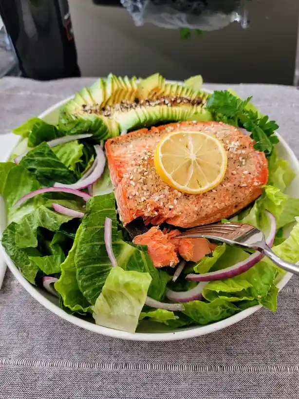 A close-up photo shows a white bowl filled with a fresh salad featuring a large piece of cooked salmon topped with a lemon…