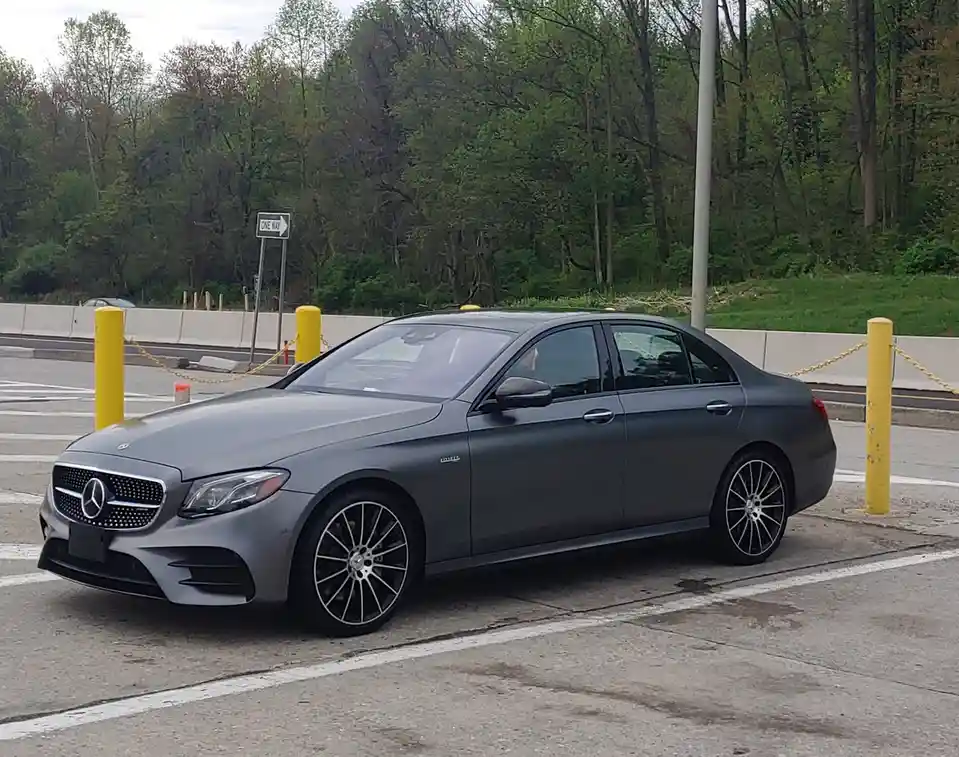 A matte gray Mercedes-Benz E63 AMG sedan is parked in a lot with white lines, flanked by yellow bollards and a chain…