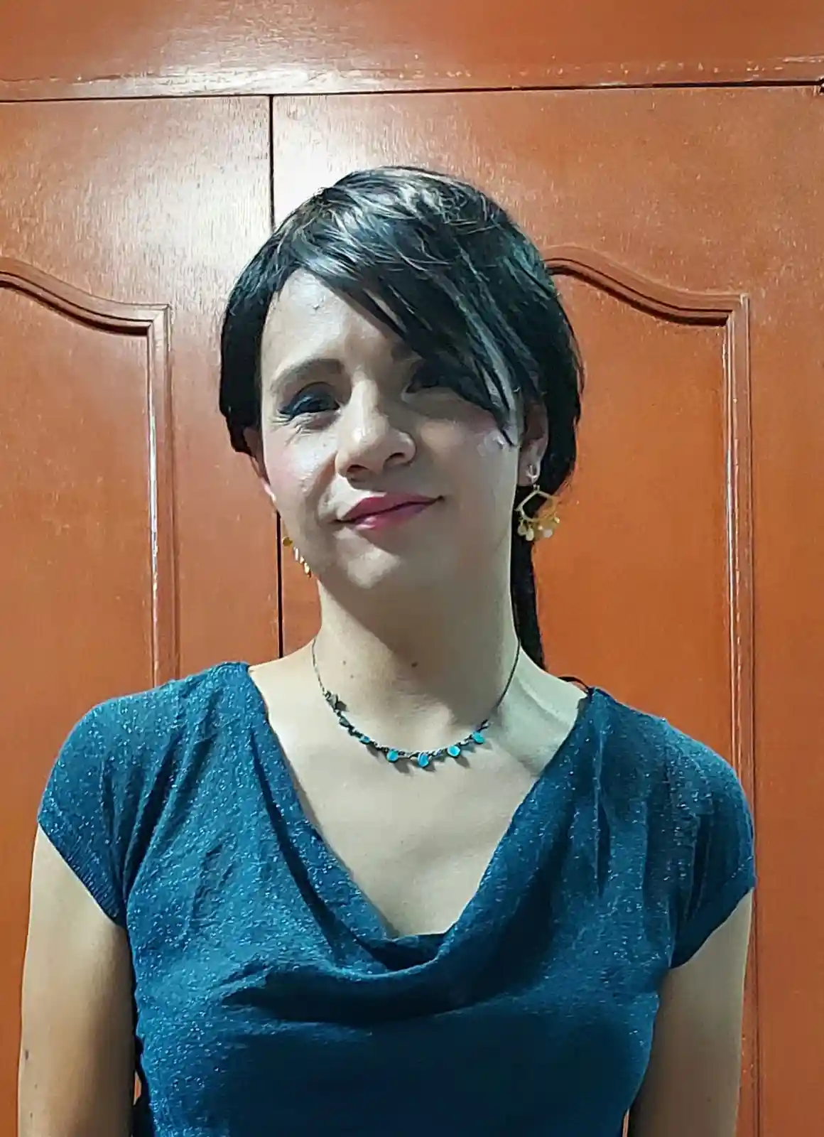 A woman with short dark hair and light makeup stands in front of a wooden door, wearing a blue top, a turquoise beaded…