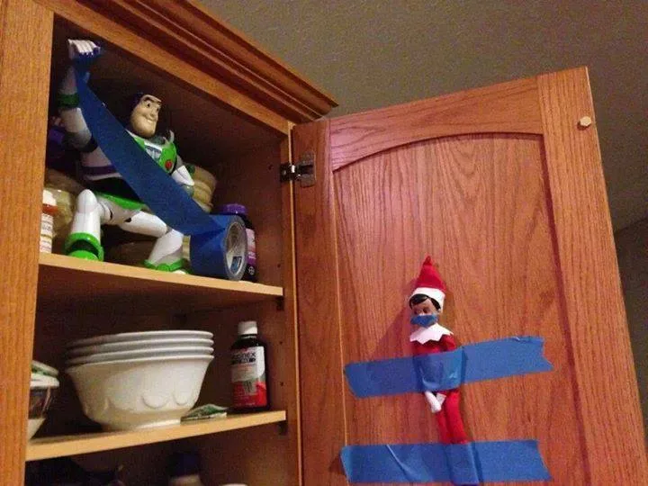 An elf on the shelf is taped to the inside of a wooden cabinet door with blue painter's tape, while a Buzz Lightyear action…