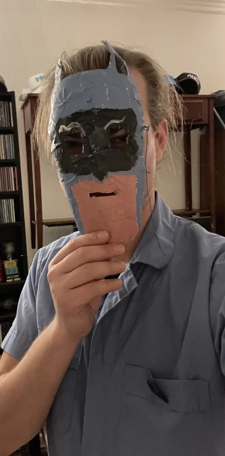 A person with light brown hair tied back is holding a crudely made Batman mask up to their face. The mask is constructed…