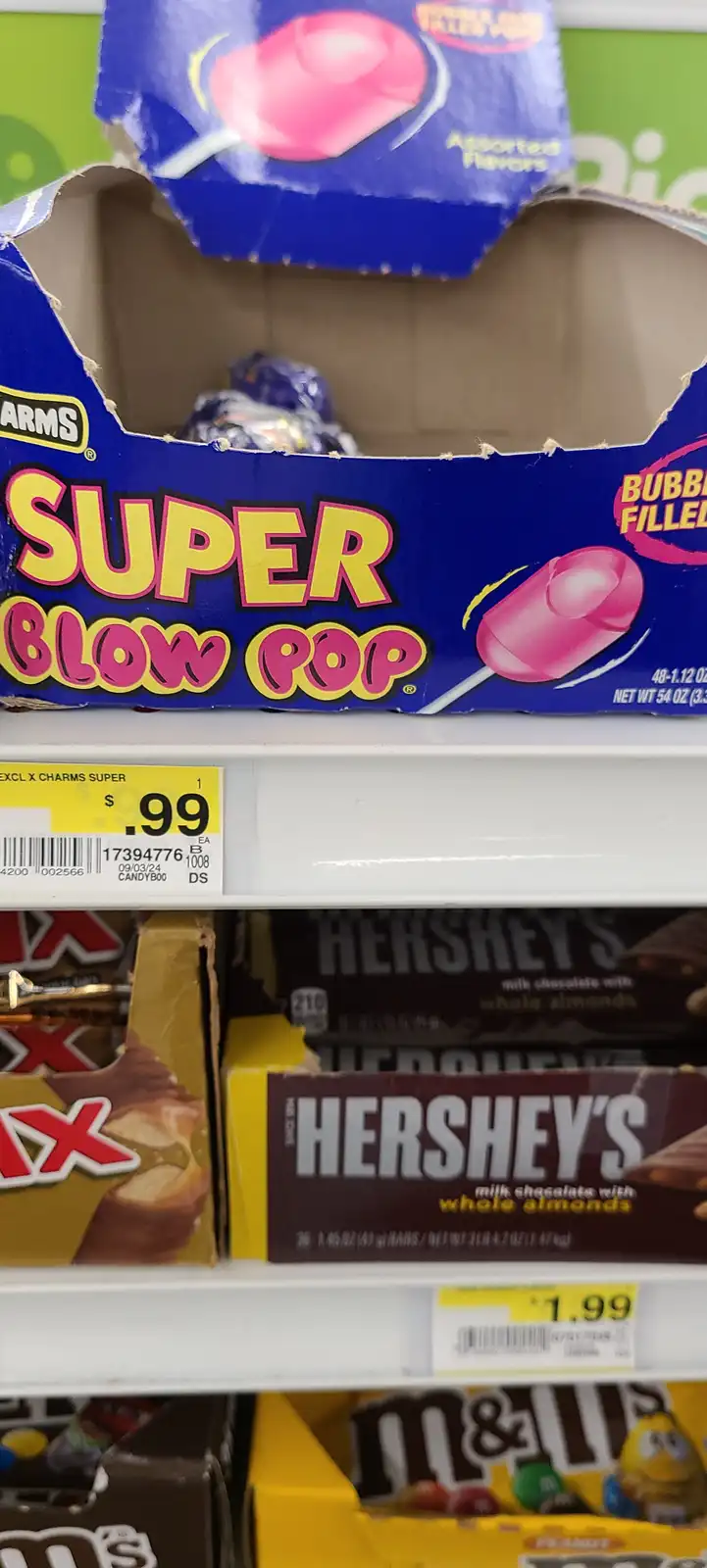 A close-up photo of a blue box of Super Blow Pop bubble-filled lollipops on a store shelf, priced at $0.99, with Hershey's…