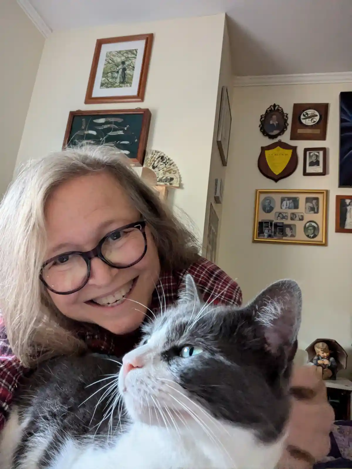 A woman with blonde hair and glasses smiles while holding a gray and white cat with bright blue eyes. The cat looks up…