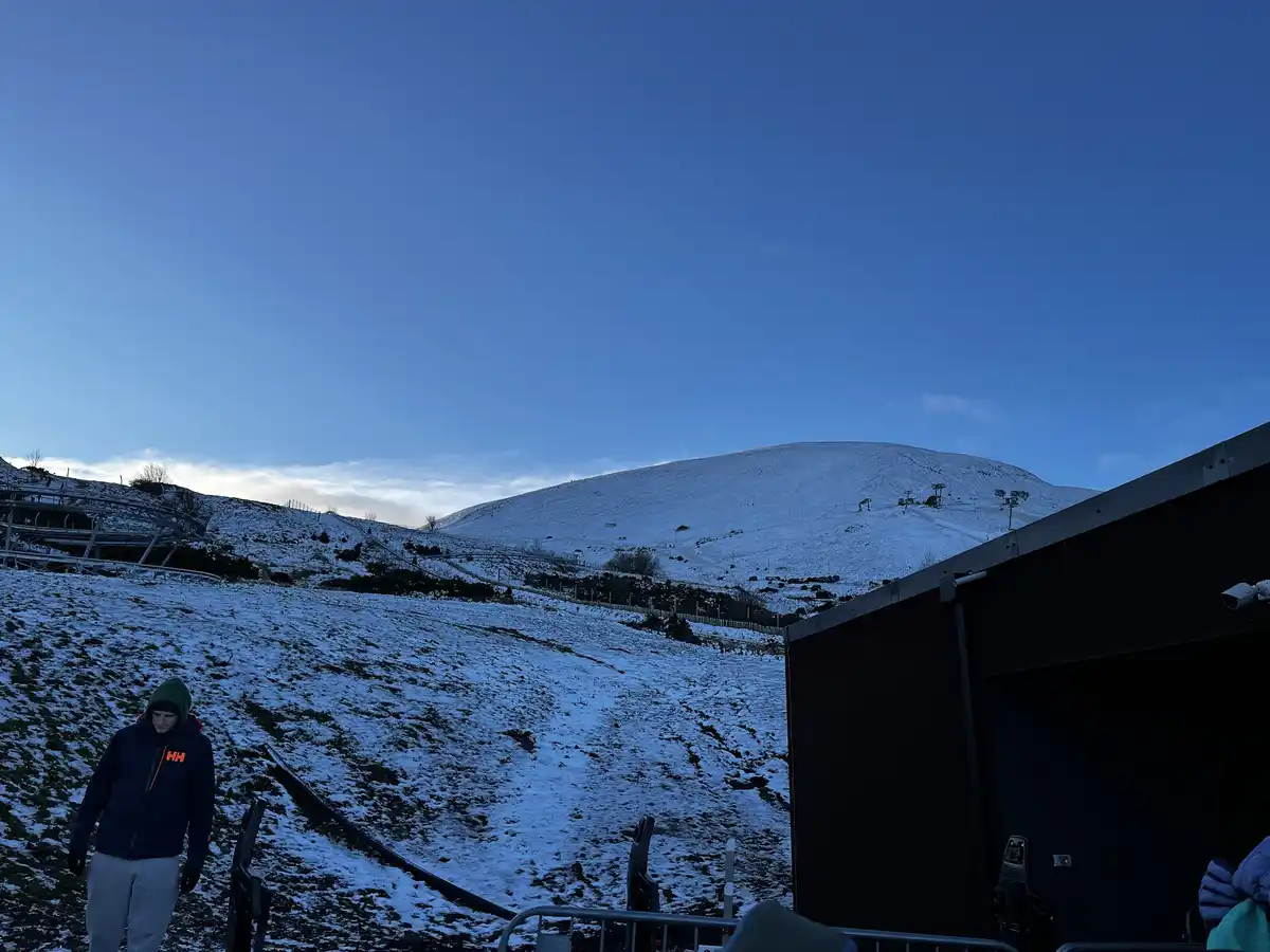 A person in winter gear stands at the base of a snow-dusted mountain with ski lifts visible in the distance under a clear…
