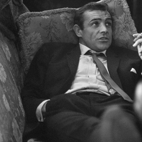 a black and white photo of a man in a suit reclining in an ornate chair, looking upward with a surprised or incredulous…