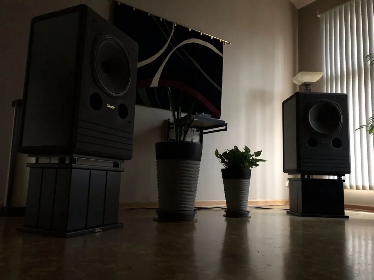 A low-angle shot of two large black Zanco floor-standing speakers on stands in a room, flanking two potted plants. Behind…