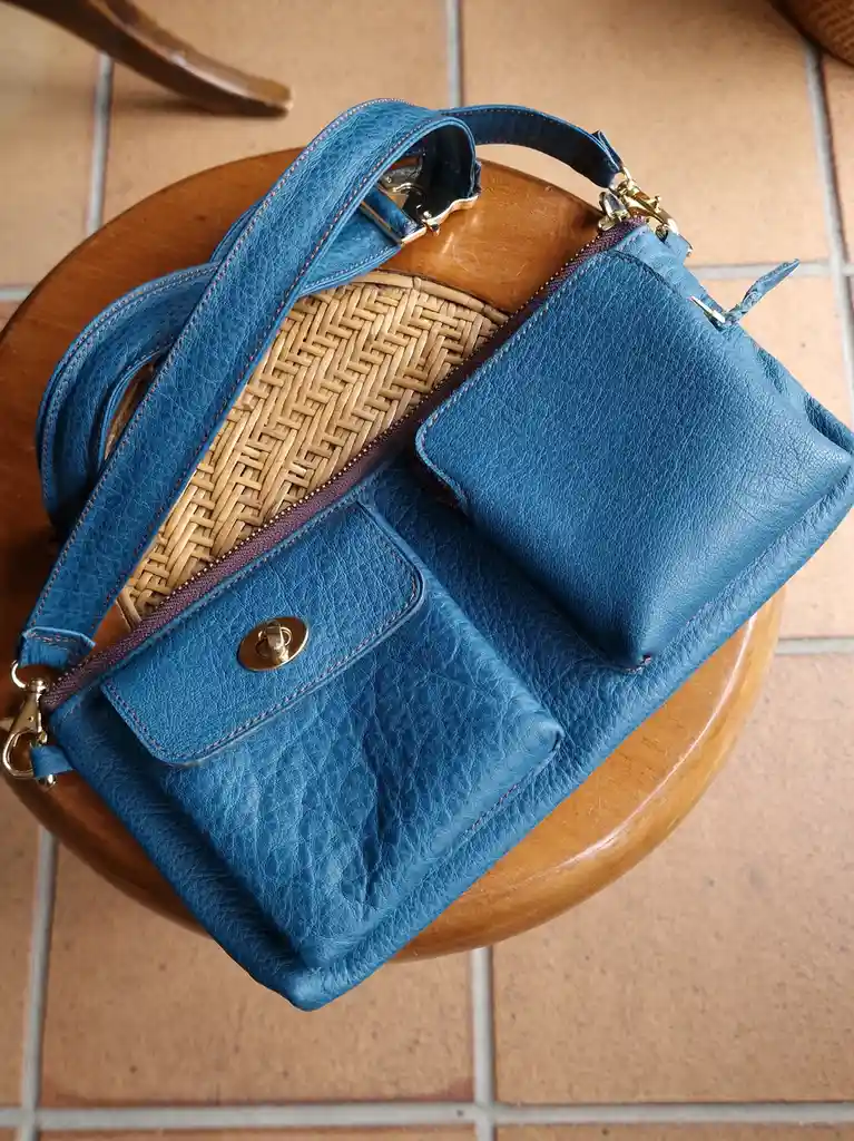 A blue leather handbag with gold hardware and a woven interior is shown open on a wooden stool, revealing its simple…