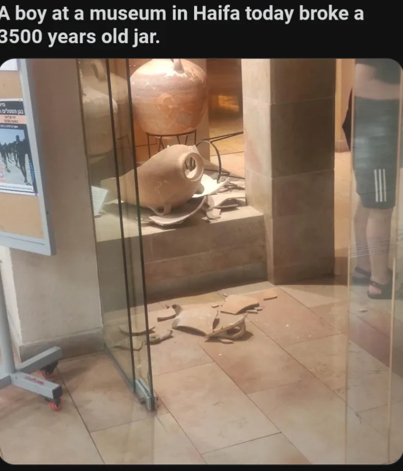 A shattered ancient clay jar lies broken on the tiled floor inside a glass display case in a museum. Shards are scattered…