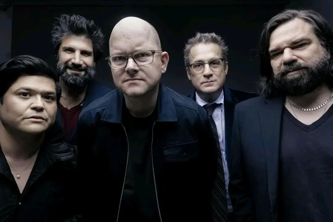 five men in dark clothing pose seriously for a group photo, with the central bald man in glasses looking intensely forward…