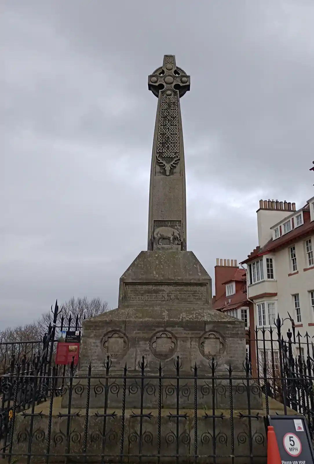 A tall stone Celtic cross war memorial, featuring carvings of a stag and an elephant, stands behind a black wrought-iron…