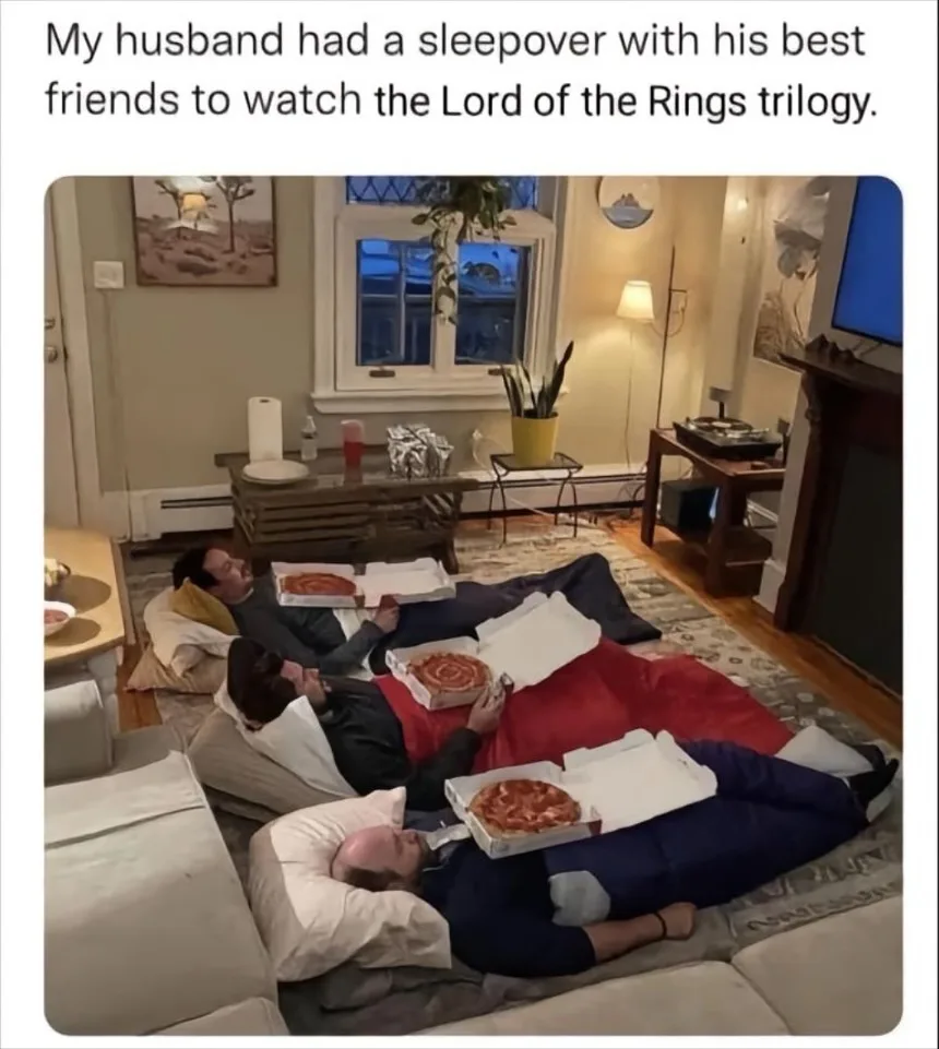 Three men are lying on the floor in a living room, each propped up on pillows with a pizza box resting directly on their…