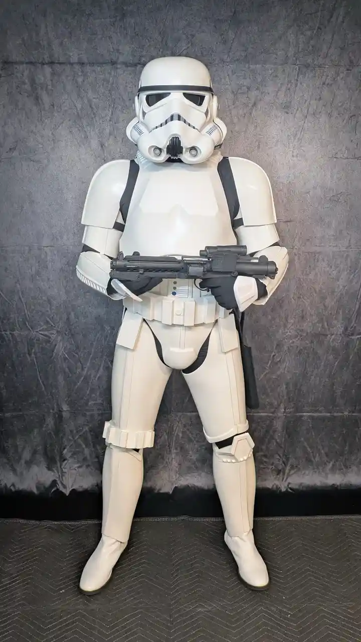 A person in a full white stormtrooper costume from Star Wars stands at attention holding a blaster rifle against a gray…