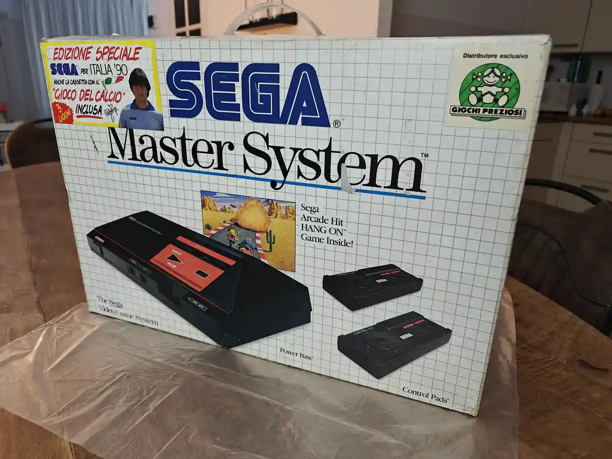 A weathered, white box for a Sega Master System video game console sits on a wooden table, showing the system, two…